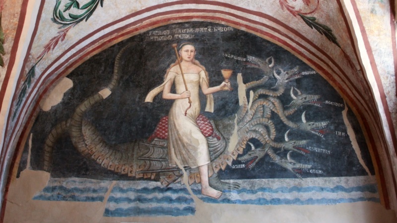 Fresco lunette on a dark blue-black ground. A young woman with long loose reddish hair, wearing a pale off-the-shoulder gown with long hanging sleeves, sits on red patterned cushions on the back of a scaly grey-green dragon. She holds a long staff in her right hand and raises a golden chalice in her left, and one bare foot rests on stylised blue and white waves. The dragon's long tail curls behind her, and seven horned heads with open, fanged mouths rise on serpentine necks to the right, each labelled with a vice: SUPERBIA, INVIDIA, ACCIDIA, IRA, AVARITIA, GULA, LUXURIA. A Latin inscription at the top names the figure as Babylon the Great. The lunette is framed by red bands, with green and red foliage on the vault above