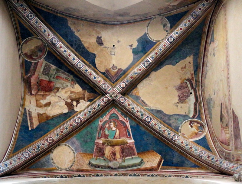 Gothic cross vault decorated with fresco, seen from below. Four diagonal ribs painted with bands of red, blue and white quatrefoil and lozenge ornament meet at a small carved boss. In the lower web, best preserved, the young Saint John the Evangelist, haloed, in a green tunic and ochre mantle, sits on a pink Gothic throne with a pointed canopy, writing at a slanting desk and raising his right hand towards his head. The throne stands on a platform with an orange patterned carpet set against a blue-green ground. The other three webs are largely lost, showing only fragments of thrones, draperies and desks on blue. Circular medallions stand at the corners of the webs; one shows a winged ox, the others are faded or empty