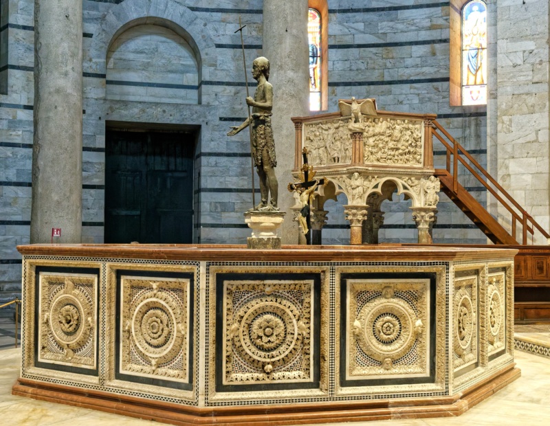 The 1246 octagonal baptismal font of Guido Bigarelli da Como at the centre of the Pisa Baptistery: sixteen marble panels with high-relief rosettes framed by Cosmatesque inlay, the bronze Saint John the Baptist by Italo Griselli at its centre, and Nicola Pisano's 1260 pulpit and the banded grey masonry of the rotunda behind