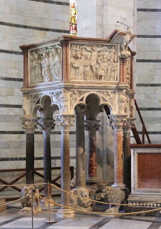 Nicola Pisano's hexagonal pulpit of 1260 in the Pisa Baptistery: seven polychrome columns, three resting on crouching lions, carry trilobed Gothic arches with Virtues and prophets at the angles; above, white marble relief panels of the Presentation in the Temple and the Crucifixion flank clustered red colonnettes, with the marble eagle lectern of Saint John at the corner beside the stair