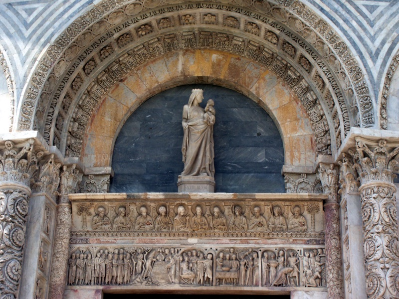 Upper portion of the east portal of the Pisa Baptistery, second half of the twelfth century: in the lunette, faced with grey-blue bardiglio, stands the white marble Madonna and Child after Giovanni Pisano; below, the marble architrave carries two relief registers — above, a hieratic procession of Christ, the Virgin, and apostles between palm trees; below, the life of Saint John the Baptist from his preaching and the Baptism of Christ to Herod's banquet and his beheading — the whole enclosed by richly carved acanthus archivolts, medallion busts, and the striped bichrome masonry of the rotunda