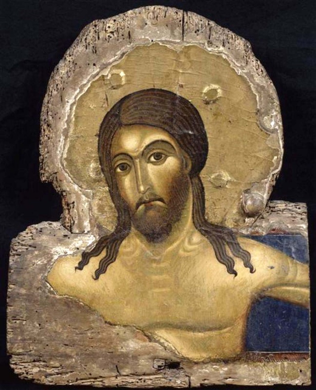 Irregular, worm-eaten fragment of a painted cross showing the head and upper chest of a living Christ. His eyes are open, his long dark hair falls in wavy strands onto his shoulders, and his bearded face is set within a raised gilded halo with empty round sockets for inlays. A strip of blue cross ground survives at the right, and the edges are broken bare wood and gesso