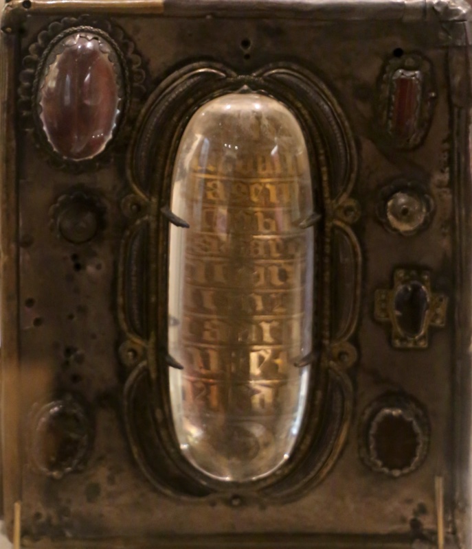 Front face of the medieval book-shrine of the Book of Mulling: a rectangular metal box with worn silver surface, dominated by a large polished oval rock crystal held by dark claw mounts at the centre; rows of gilt Gothic lettering on silver foil are visible magnified through the crystal. Around it are several smaller settings — dark red and brown oval polished stones in decorated mounts, a small cross-shaped fitting and plain bosses — framed by twisted rope-like mouldings, with dents and losses at the corners. The shrine stands on a pale surface in a museum display