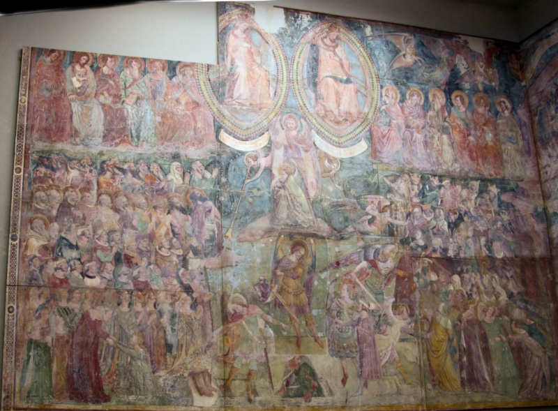 Large medieval fresco of the Last Judgement. At the top, Christ and the Virgin Mary sit side by side in two almond-shaped glories with rainbow borders, flanked by rows of seated apostles. Below, angels blow trumpets and hold scrolls, while armoured archangels with swords separate two dense crowds of resurrected figures: the blessed on the left, the condemned on the right. The painted surface is faded and shows the joins of the detached sections.