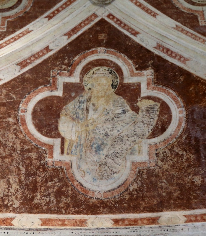 Gothic vault web frescoed in mottled red-brown, framed by white ribs with red ornament. A quatrefoil medallion encloses a haloed bearded figure whose halo has turned black, draped in pale blue-grey robes, holding up a large unfurled scroll; the painted surface is severely abraded, with the design surviving mainly as ghostly outlines