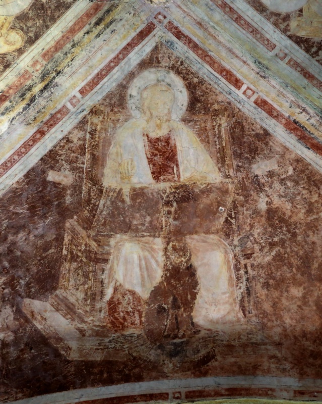 Gothic vault web in mottled red-brown framed by white ribs with red ornament. A haloed, bearded evangelist in a dark red tunic and pale mantle sits frontally on a wide architectural throne, one hand raised before his chest; at his feet, between the folds of his robe, crouches a dark, heavily damaged four-legged winged creature, his evangelist symbol. The surface is abraded throughout