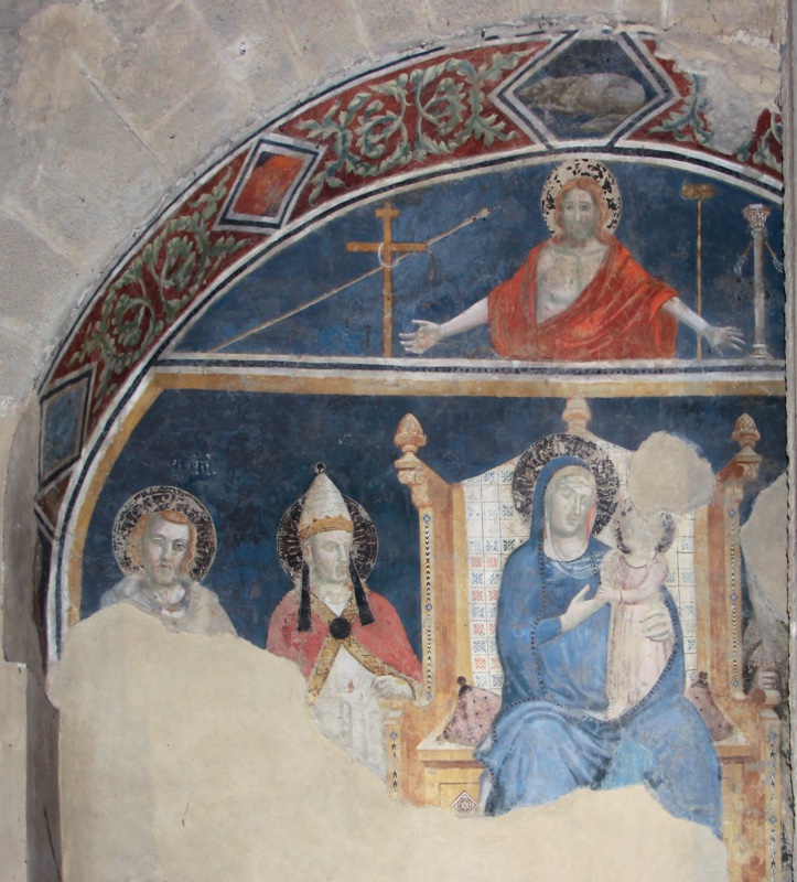 Fragmentary medieval fresco under a stone arch. In the lunette above, a half-length Christ with open arms displays his wounds among the instruments of the Passion against dark blue. Below, the Virgin in a blue mantle sits on a golden throne with a patterned cloth of honour, holding the standing Christ Child whose face is lost; to the left stand a young saint and a sainted pope in a white tiara and red cope. Large areas of bare plaster interrupt the scene