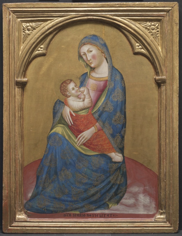 Gold-ground panel under a round gilded arch with carved foliage in the corners. The Virgin, in a blue mantle with large gold motifs and a green lining over a pink dress, sits low on a broad red ground and nurses the Child, who wears a red tunic patterned in gold. A small gold sun with rays shines on her breast and a gold star on her shoulder, faint gold rays spread around her, and a Latin inscription naming the Madonna of Humility runs along the bottom