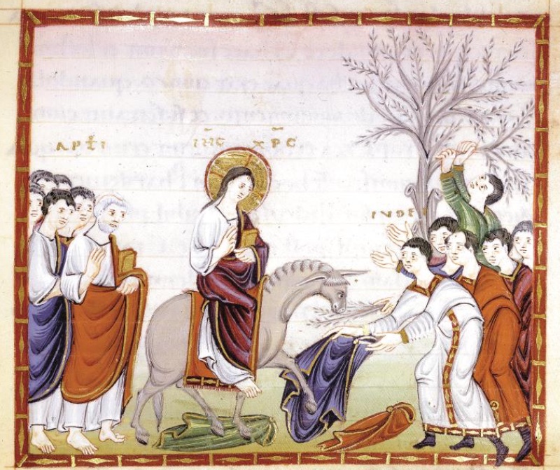 Medieval miniature in a red frame with gold ornament, on a pale ground with faint show-through writing: at left a group of apostles, the foremost white-haired in a red mantle holding a book; at centre a haloed Christ in a white tunic and red-brown mantle rides sideways on a grey ass, holding a book and raising his hand; green, blue and red garments lie on the ground before the animal, which lowers its head towards a branch held out by a young man; at right a crowd of beardless youths in white tunics and red mantles stretch out their hands, while one in green climbs a grey tree to break off a branch