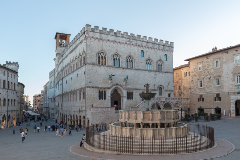 Two-tiered polygonal marble fountain with carved reliefs, dusted with snow, standing before the crenellated Gothic façade of the Palazzo dei Priori in Perugia.