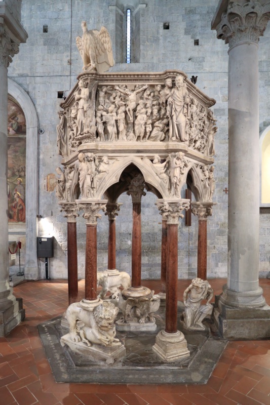 Hexagonal white marble pulpit on slender columns, its parapet carved with five deeply undercut relief panels of crowded figures; lions and a crouching male figure support the columns at the base.