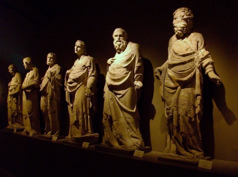 Eight over-lifesize marble figures standing in a receding rank on a low plinth against a dark wall, lit from the right: the two nearest are pale and comparatively well preserved, while those behind are discoloured, deeply pitted and abraded. All lean forward from the hips with necks extended and heads carried in advance of the chest.