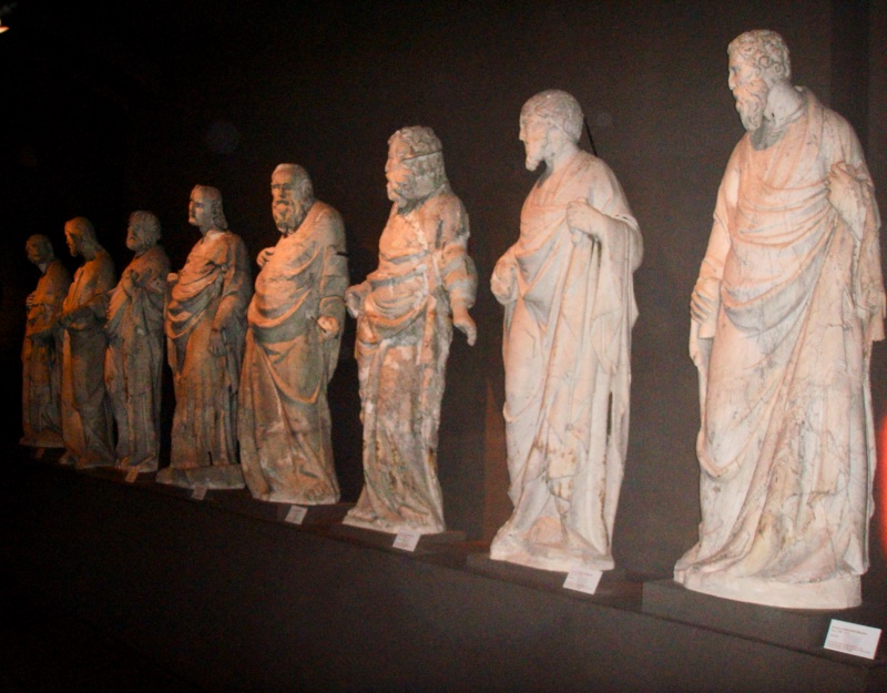 Eight over-lifesize weathered marble figures standing in a receding rank against a dark ground, lit obliquely from above: bearded and youthful men in heavy classical drapery, their necks long and heads thrust forward, several leaning markedly out from the hips.