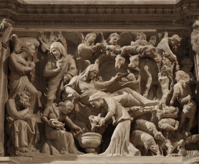 Deeply undercut marble relief: at the upper left a winged angel addresses a standing veiled woman, with a bearded man seated in thought below; across the centre a veiled Virgin reclines and reaches toward a swaddled infant in a manger, the heads of an ox and an ass leaning above; at the right angels and shepherds stand among trees with sheep and goats below; at the lower centre a woman pours water into a large footed basin while a seated woman holds a naked infant.