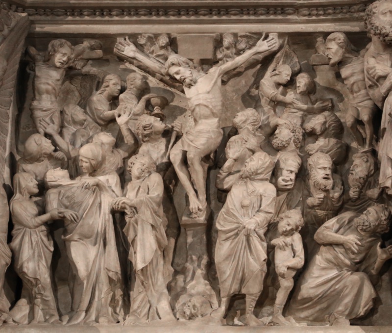 Deeply undercut marble relief: the emaciated body of the crucified Christ at the centre, head fallen forward, feet set one above the other on a small ledge with a skull beneath; at the left a veiled woman collapses backward into the arms of her companions while a draped figure stands with bowed head beside the cross; below, a soldier drives a lance upward with both hands; at the right a dense crowd of bearded and youthful faces looks up, and an angel at the upper corner pushes a small nude figure downward.