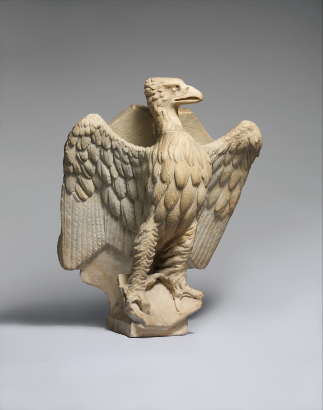 White marble eagle standing with wings raised and spread, head erect and turned to one side, talons grasping a book; a hexagonal bookrest with a projecting lower ledge rests across the raised wings. The plumage is carved in overlapping scale-like feathers on the breast and long striated flight feathers at the wing edges.