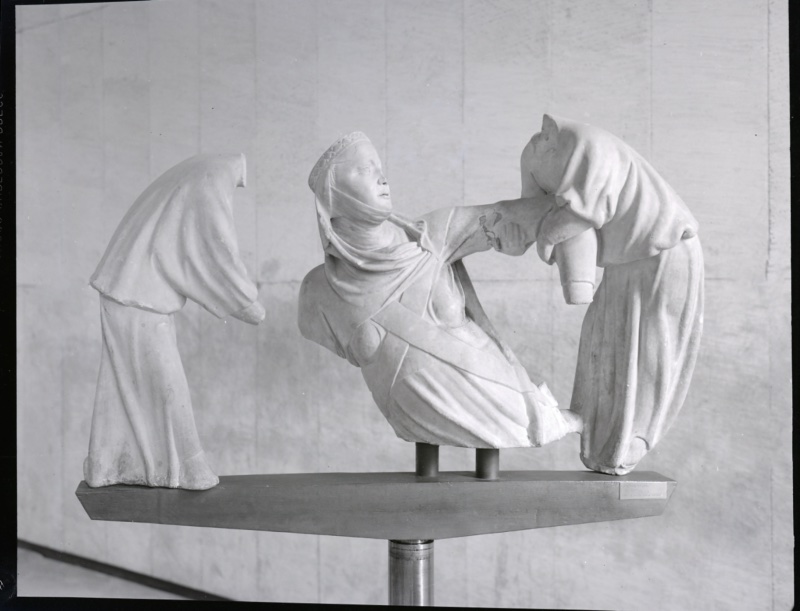 Black-and-white photograph of three marble figures on a metal display mount: at the centre a woman in a pierced coif and wimple, head tilted back and eyes lifted, rising from a mass of drapery; at either side a figure bent steeply forward, face hidden within a deep hood, arms extended toward her.