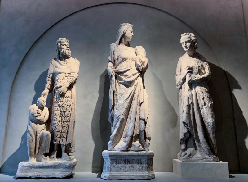 Three weathered marble figures standing in a shallow vaulted niche: at the left a gaunt bearded man in a shaggy fleece garment rests his hand on the head of a small kneeling figure with joined hands; at the centre a crowned and veiled woman on an inscribed plinth holds a headless child on her left arm; at the right a youthful figure holds a book against his chest.