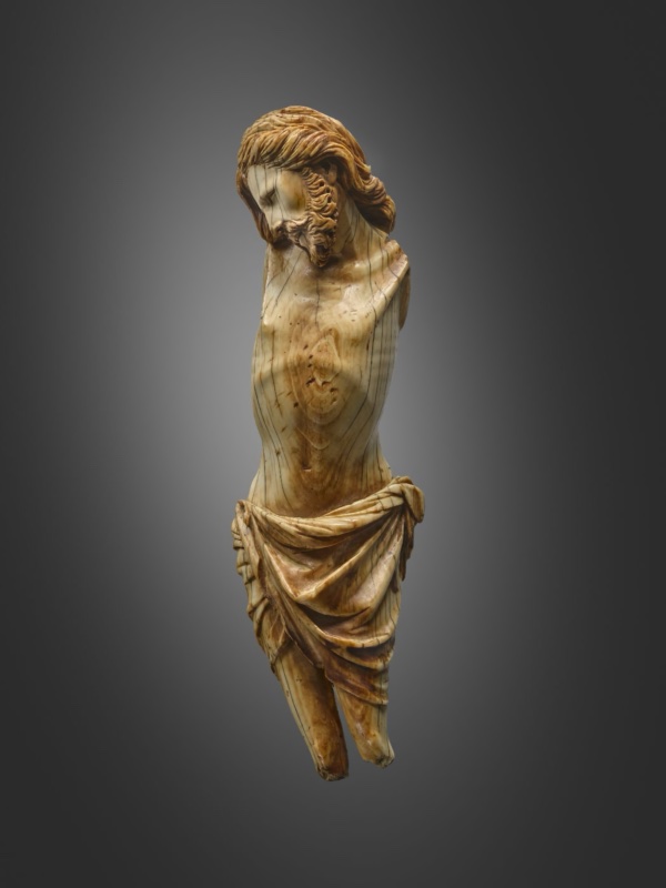 Small ivory figure of the crucified Christ, gold in colour with the head falling onto the right shoulder with eyes open beneath contracted brows, long striated hair framing a curled beard; the torso is attenuated with the ribcage sharply articulated, and a knotted loincloth falls in deep folds. The arms are broken away at the shoulders and the legs below the calf.