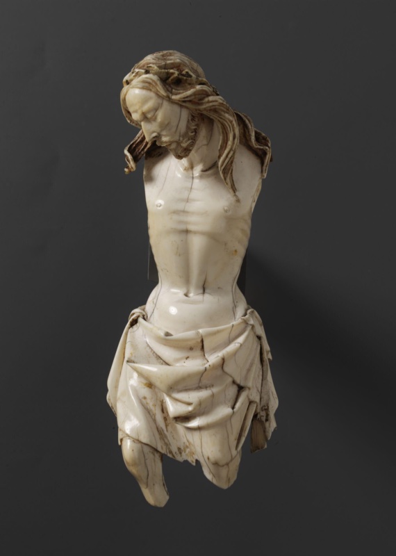 Small ivory figure of the crucified Christ photographed against a grey ground: the head falls forward and to one side with eyes closed, long waved hair and a divided beard retaining traces of gilding; the torso is carved with sharply defined clavicles, ribs and abdominal channel; a loincloth knotted at the hip falls in broad planes broken by angular projecting folds. The arms are broken away at the shoulders and the legs below the knee.