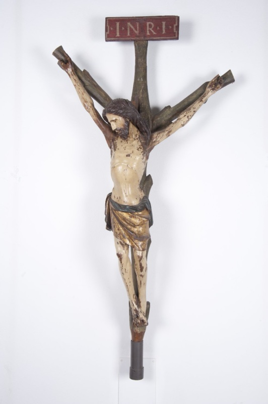 Polychrome wooden crucifix on a white wall: the emaciated body of Christ hangs with arms stretched steeply upward, head fallen forward and to one side with eyes closed, dark waved hair and divided beard; the loincloth is gilded with a green border, and the cream flesh shows extensive paint loss exposing a brown ground. The plain cross carries a red tablet lettered INRI, and the shaft ends in a metal socket.
