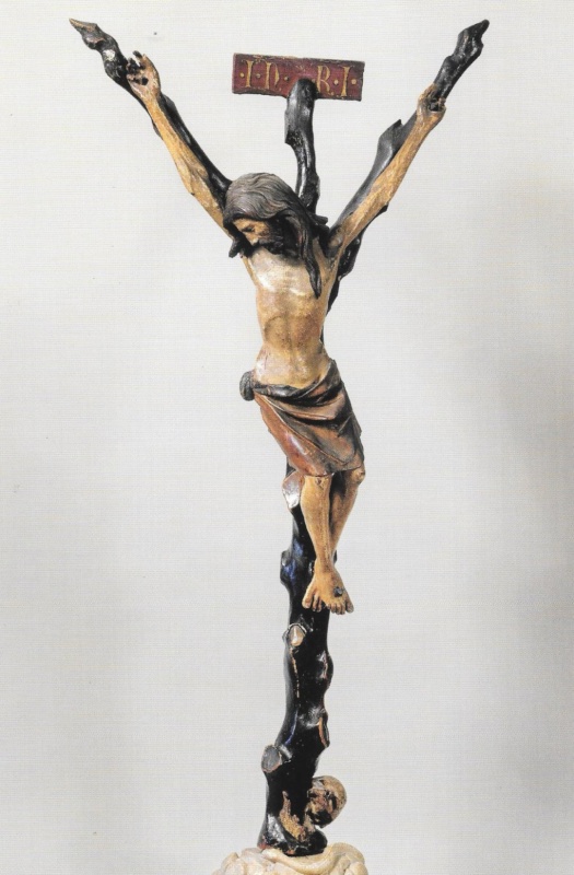 Polychromed wooden crucifix against a pale ground: the body of Christ hangs with arms stretched steeply upward, head fallen onto the right shoulder with eyes closed, long dark hair and a divided beard; the loincloth is reddish-brown with gilded edges. The cross is carved as a dark tree trunk with the stubs of cut branches, rising from a rocky mound at whose foot lies a skull, with a red titulus lettered in gold above the head.