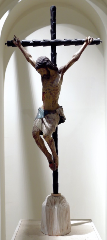 Painted wooden crucifix set in a shallow white niche on a modern conical base: the body of Christ hangs with arms stretched taut, head fallen forward, the chest pulled upward and the abdomen hollowed; blood runs from the wound in the right side; the knees bend sharply so that the legs swing to one side, the feet set one above the other. The cross is a dark stripped trunk with evenly spaced branch-stubs along shaft and arms.