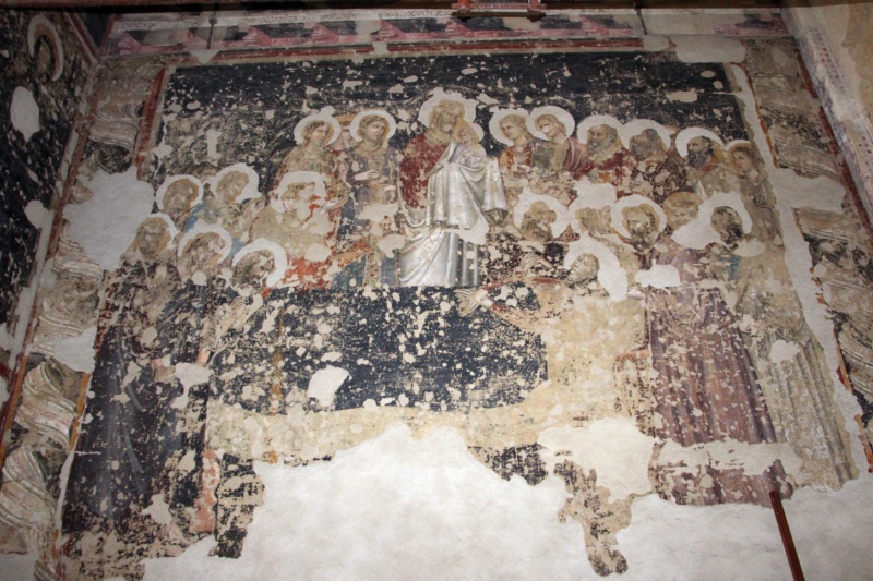 Damaged fresco of the Dormition of the Virgin by Giovanni da Rimini in the church of Sant'Agostino, Rimini. At the centre Christ, in a red mantle, stands behind the deathbed holding the Virgin's soul as a small figure wrapped in white. Crowds of haloed apostles and young figures surround him in overlapping rows. The Virgin lies on a long dark bier, her pale face visible at the right end. The dark background has lost most of its blue paint, large patches of plaster are missing, and painted twisted columns frame the scene at both sides