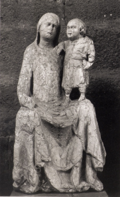 Weathered marble statue attributed to Giovanni di Agostino, c. 1331, from a dismembered saint's shrine in Volterra: a seated, veiled Madonna with heavily eroded features inclines her head toward the Christ Child, who stands upright on her knee in a belted tunic, reaching one hand toward her face; photographed against rough masonry; Museo Diocesano d'Arte Sacra, Volterra.