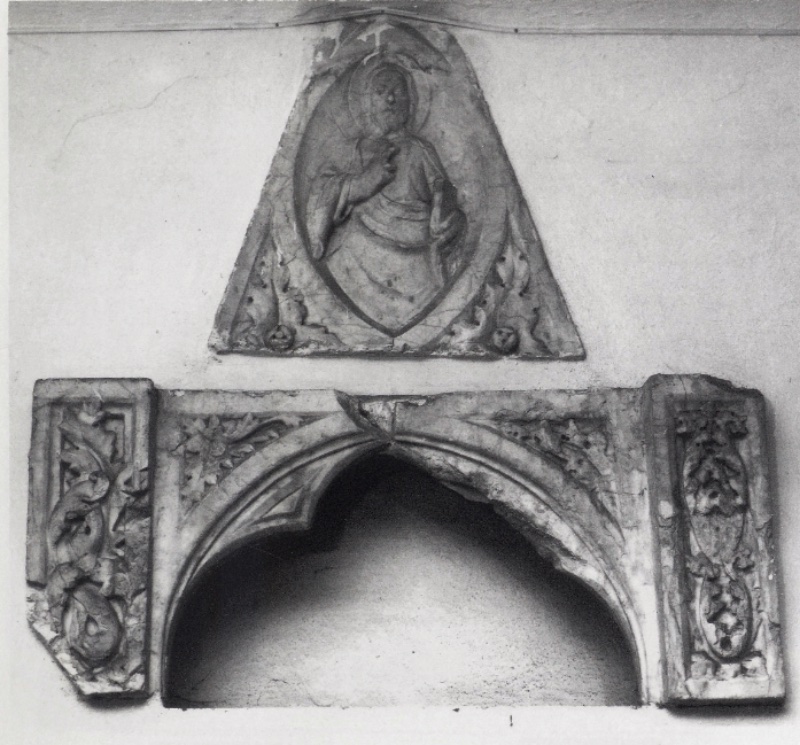 Two marble fragments of a tabernacle attributed to Giovanni di Agostino, c. 1331, set into a plastered museum wall: above, a triangular gable with the half-figure of Christ blessing in a mandorla, cross-nimbed, a book at the frame; below, a cusped ogee-arched niche opening into a dark cavity, flanked by pilaster strips of scrolling vine; Museo Diocesano d'Arte Sacra, Volterra