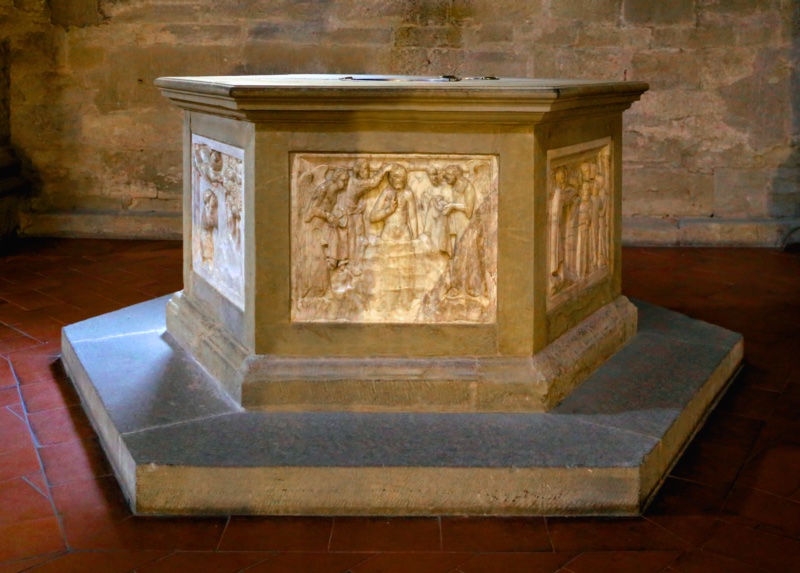 Hexagonal baptismal font in the Pieve di Santa Maria, Arezzo, on a stepped stone base: three white marble relief panels attributed to Giovanni di Agostino, c. 1332, set into its faces — the Baptism of Christ at the centre between the young John led into the desert and the Ecce Agnus Dei — beneath a plain moulded cornice.