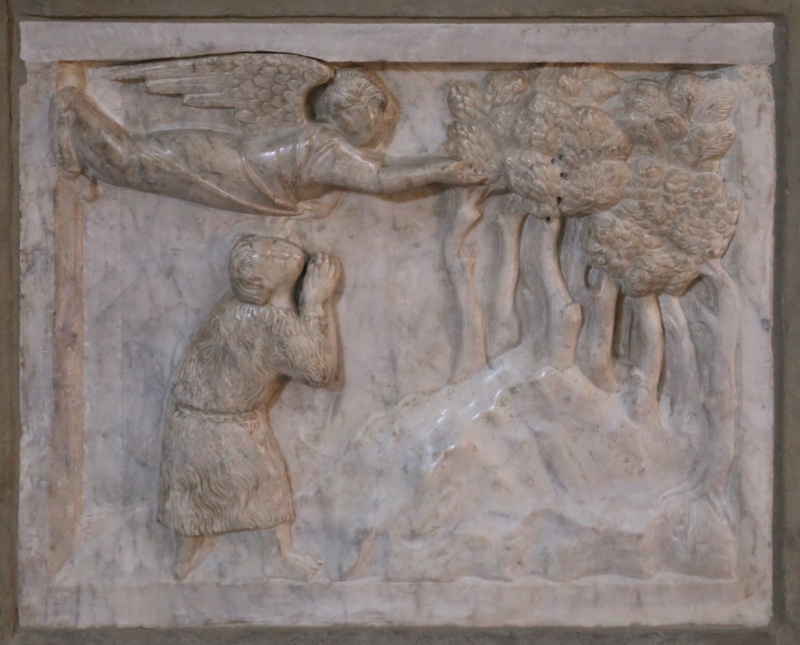 Marble relief attributed to Giovanni di Agostino, c. 1332, on the baptismal font of the Pieve di Santa Maria, Arezzo: an angel flies horizontally across the top, drapery streaming over the frame's edge, arms extended toward a grove of trees on a smooth rocky hill, while below the young Baptist in his tufted camel-hair tunic climbs with hands joined in prayer, face upturned to the messenger