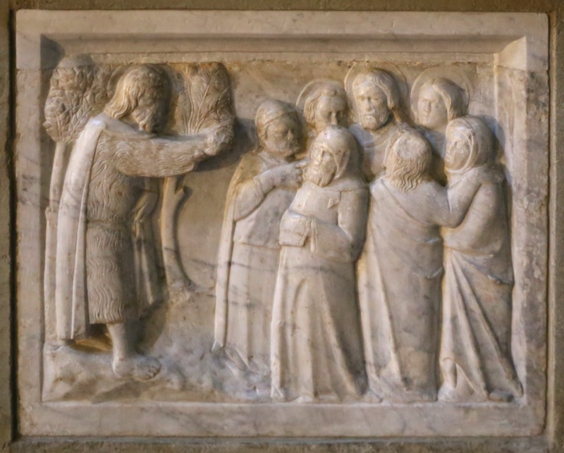 Marble relief attributed to Giovanni di Agostino, c. 1332, on the baptismal font of the Pieve di Santa Maria, Arezzo: the Baptist in his camel-hair tunic stands at left among trees, arm extended toward a pressed crowd of seven figures at right, within which the haloed Christ gazes frontally at the viewer between two haloed companions