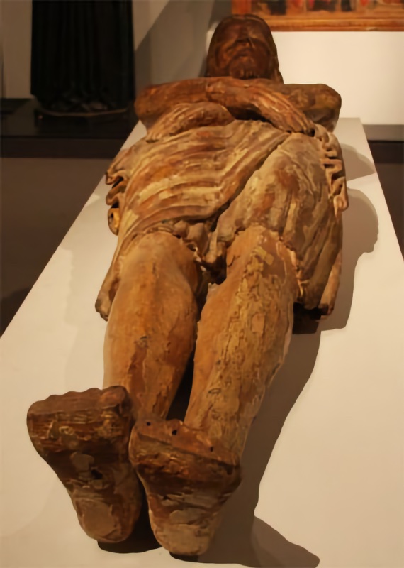 Life-size wooden statue of the dead Christ lying on his back, seen foreshortened from the feet, whose soles show carved nail holes. The forearms are crossed over the lower chest, the bearded head rests with closed eyes, and a deeply folded cloth wraps the hips. The painted surface is almost entirely worn away, exposing bare brown wood