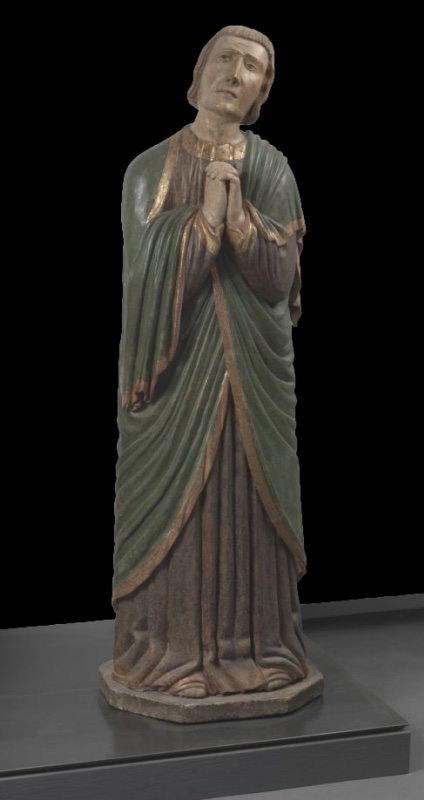 Polychrome wooden statue of a young, beardless St John the Evangelist standing with hands tightly clasped at his chest. His head tilts toward one shoulder, brows knotted and eyes raised in grief. He wears a grey-brown tunic with a gilded collar under a deep green, gold-bordered mantle that crosses the body in heavy diagonal folds, falling to his feet on a small octagonal base