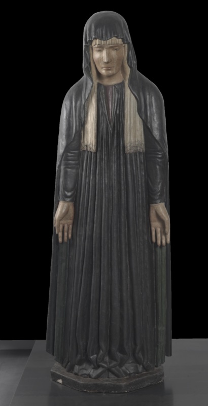 Polychrome wooden statue of the grieving Virgin standing upright, entirely wrapped in a dark hooded mantle falling to the ground. A white veil with ornamented ends frames her pale, still face with lowered eyes, and descends over a dark violet dress carved in dense vertical pleats. Her bare hands hang open and empty at her sides in a gesture of helpless sorrow