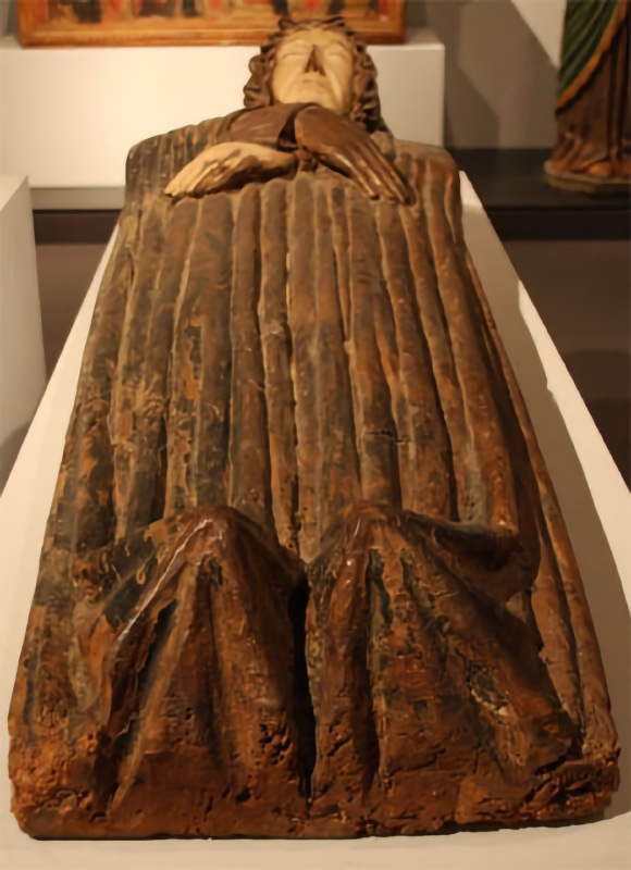 Life-size wooden effigy of the dead Virgin lying on her back, seen foreshortened from the feet. Her forearms are crossed over the breast, one hand laid over the other arm, her pale aged face rests with closed eyes, framed by carved waves of hair. The dress covering the whole body is carved into long, thin parallel pleats that break over the feet in sharp angular folds. The painted surface is largely worn away to bare brown wood