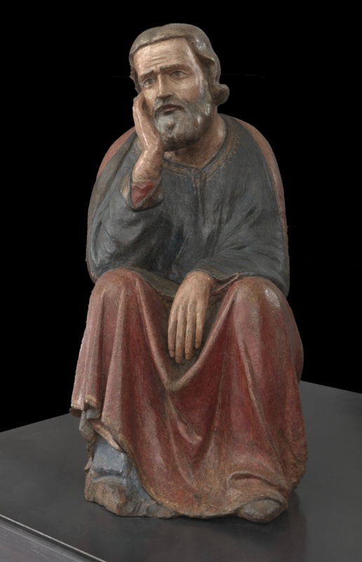 Fourteenth-century painted wooden statue of St Joseph seated on a rock, resting his bearded cheek on his right hand in a melancholy pose; he wears a dark blue-grey tunic with gilded borders under a red mantle draped over his knees. Maestro dei Magi di Fabriano, Pinacoteca Civica Bruno Molajoli, Fabriano.