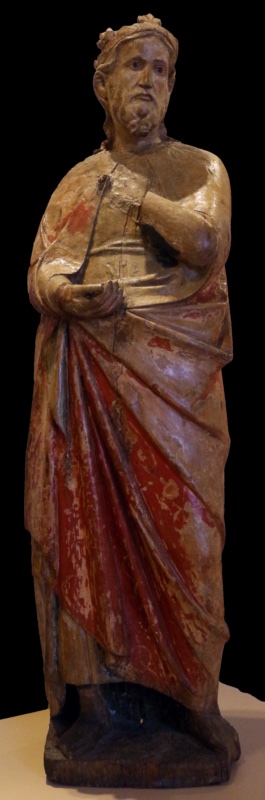 Fourteenth-century life-size painted wooden statue of a bearded, crowned Magus with lowered head and pensive expression, a slender dark fragment lying across his open right hand, where his gift is lost; his cinnabar-red mantle with traces of gilding falls in long diagonal folds. Maestro dei Magi di Fabriano, Museo Nazionale del Palazzo di Venezia, Rome.