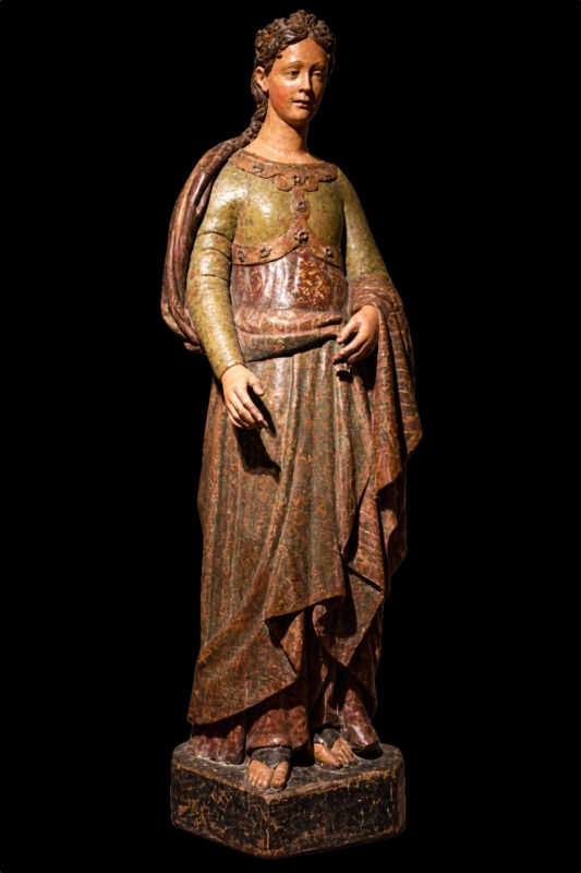 Fourteenth-century life-size painted wooden statue of a standing woman, presented in 2014 as St Helena though no surviving sign confirms it, with braided hair coiled about her head and no crown, in a fitted green bodice with applied bosses and a patterned skirt and mantle; a dark stump of the lost attribute survives between the fingers of her left hand at the hip. Attributed to the Maestro dei Magi di Fabriano, private collection.