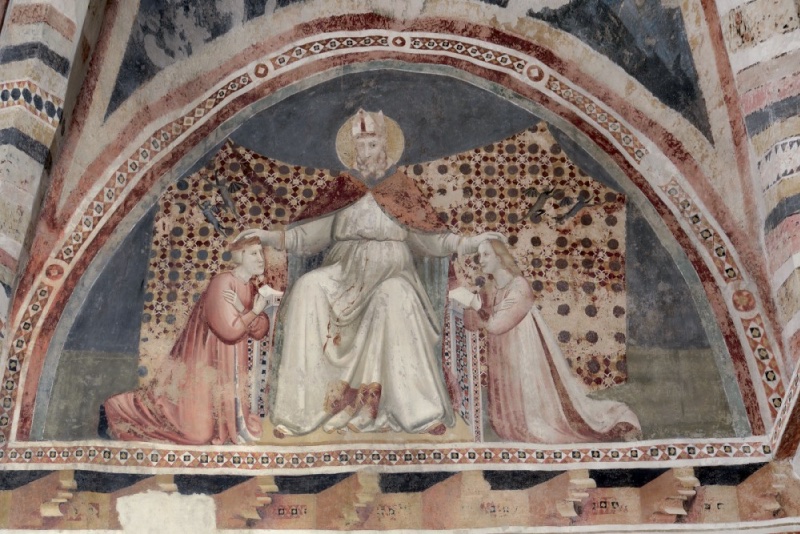 Fourteenth-century fresco lunette: a mitred bishop saint, enthroned frontally in a red cope over white robes, extends both arms to place his hands on the heads of two kneeling young figures in courtly dress, each holding a small white object. Two differently patterned cloths of honour hang behind; small dark winged creatures — the expelled demons — fly toward the upper corners. A painted corbel cornice runs below; ornamental borders frame the arch.