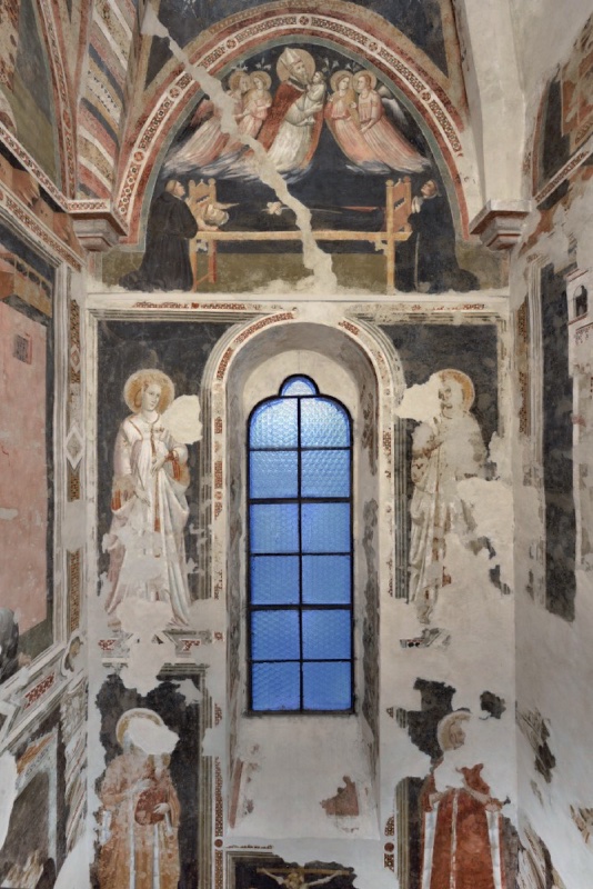 Gothic chapel wall in fresco: a pointed lancet window with blue leaded glass is framed by painted marble ornament and four standing saints on two levels, several damaged. In the lunette above, a dead monk lies on a bed mourned by black-robed friars, while a mitred bishop amid four angels carries the small figure of his soul heavenward