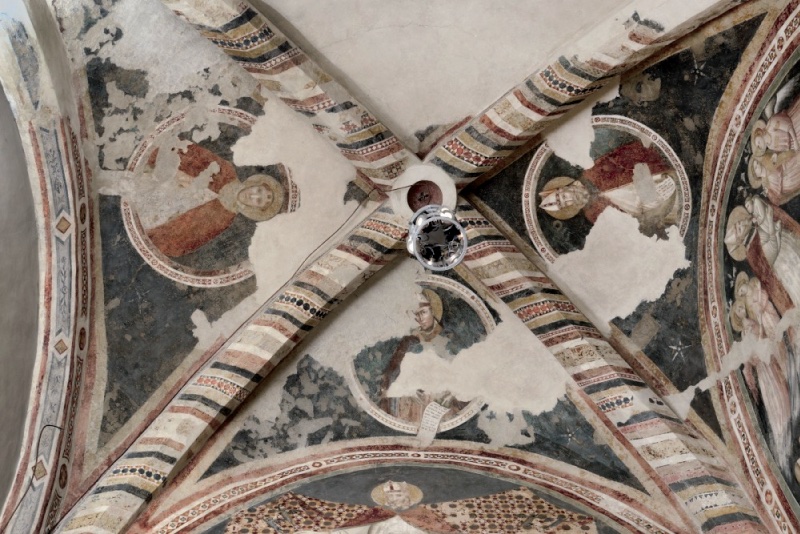 View up into a quadripartite vault: four ribs painted with bands of chevrons, chequers and rosettes meet at a central boss. In the dark blue-grey webs, roundels contain half-length saints — a figure in a red mantle, a mitred bishop with a book, and another bishop holding an open scroll — with large areas of plaster loss