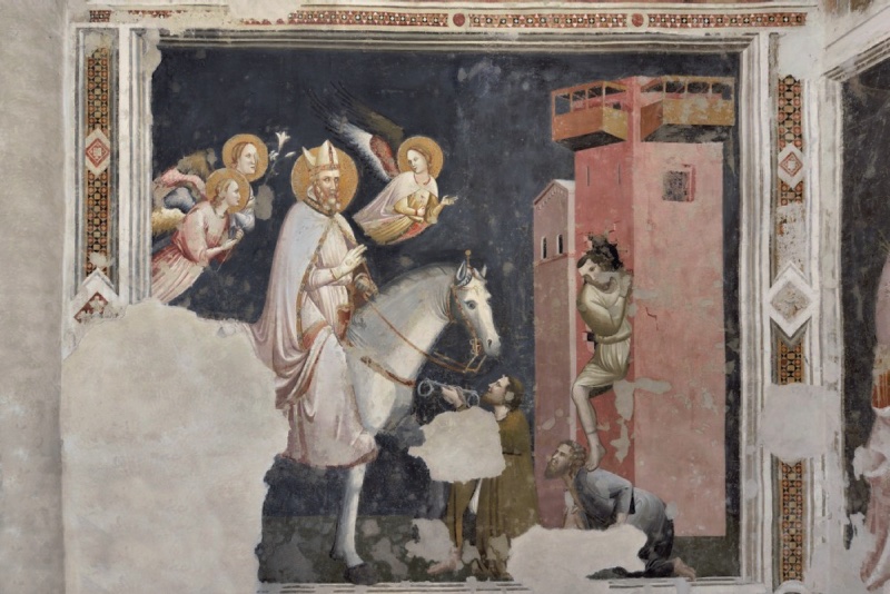 Fourteenth-century fresco on a dark blue ground: at left a haloed bishop saint rides a white horse, right hand raised in blessing, attended by three angels, two holding lilies and one flying above. At right a tall pink prison tower with a wooden gallery; a man in a pale tunic climbs down from an opening, stepping on a crouching companion, while another figure holds up broken fetters toward the saint. Large plaster losses interrupt the lower centre.