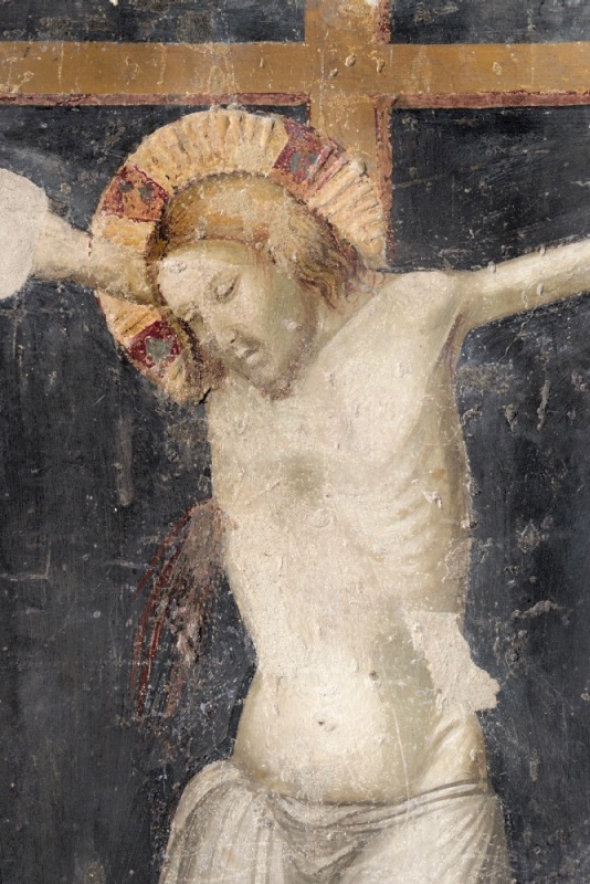 Close view of a fourteenth-century fresco Crucifixion: the head and torso of Christ on an ochre cross against a dark slate-blue ground. His head, in a radiate halo with red jewelled compartments, sinks to the right shoulder with closed eyes; thin rivulets of dark red blood run from the side wound down his flank; a transparent loincloth veils the hips. The pale surface is worn and scratched