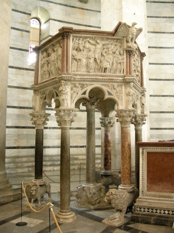 Hexagonal marble pulpit raised on granite columns, three of them standing on carved lions; round-headed trefoil arches span the supports, and the parapet above carries deeply carved relief panels of crowded figures, with an eagle lectern projecting at the upper right.