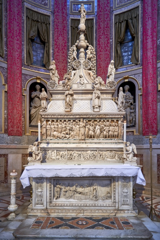 Tiered white marble funerary monument standing on an altar within a richly hung chapel: a densely carved narrative sarcophagus at its centre, a scaled lid above bearing standing saints, and a tall pinnacle crowned by a blessing figure; two kneeling angels holding candlesticks flank the base.