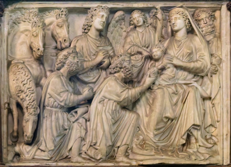 High-relief marble panel: three horses crowd the left edge, their heads foreshortened; at the centre the Magi press forward, two kneeling with bowed heads and one standing; at the right the veiled Virgin sits enthroned with the Child leaning forward from her lap, a bearded Joseph at the far edge.