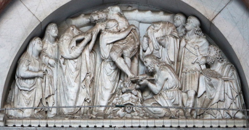 Weathered marble relief filling a semicircular lunette: the lifeless body of Christ slumps diagonally from the cross into the arms of a bearded man who takes its weight; at the lower right another kneels and works at the feet with pincers; at the left a veiled woman draws the released hand to her cheek, with a file of veiled mourners behind her.