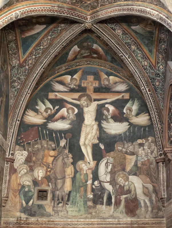 Fresco of the Crucifixion filling a pointed lunette beneath a ribbed vault. Christ hangs on a tall cross inscribed INRI, flanked by six grieving angels whose bodies trail into clouds. On the left, the holy women support the fainting Virgin beside a small barred window cut into the wall; soldiers with spears, a sponge on a reed and a red banner crowd behind a rider on a brown horse. Mary Magdalene in green embraces the foot of the cross, beside a small kneeling donor in black. On the right, a rider on a white horse and helmeted soldiers look up at Christ, one raising his hand, while Saint John sits mourning in the lower corner. Ornamental bands and Evangelist symbols decorate the vault above